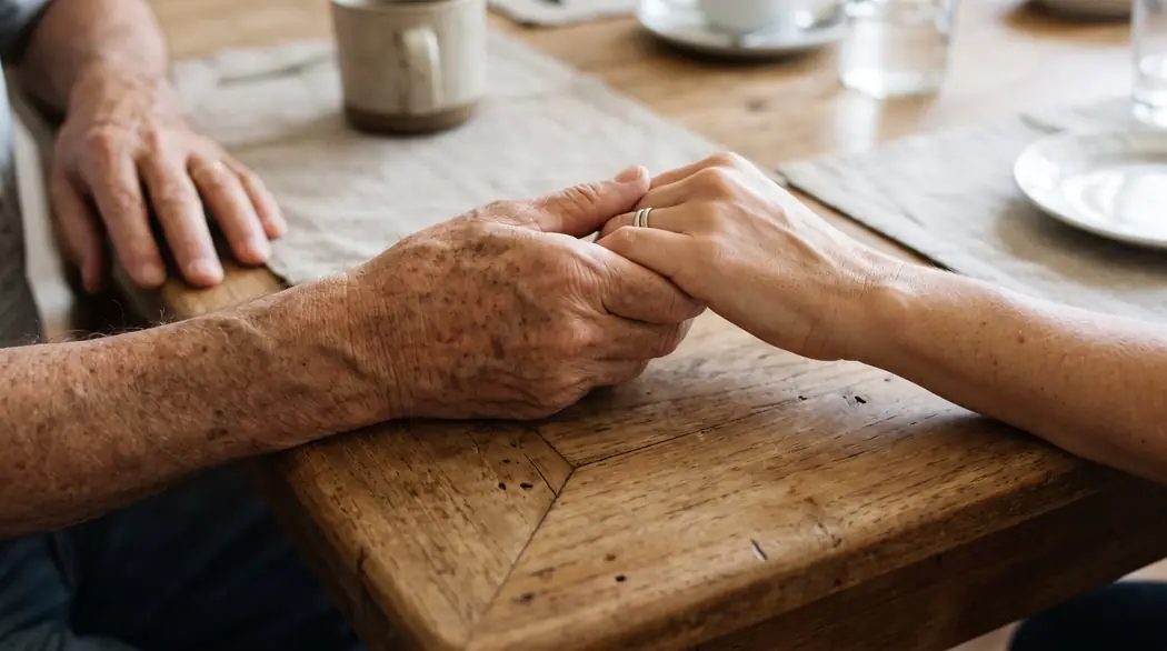 Mano de un hombre mayor sosteniendo con cariño la mano de su hija sobre la mesa